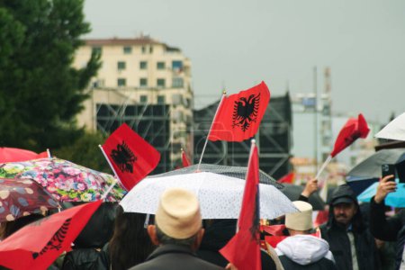 Foto de Tirana, Albania - 28 de noviembre de 2023: A lo largo de la Avenida de los Mártires de la Nación, la gente participa alegremente en una marcha del Día de la Independencia en un día lluvioso, sosteniendo banderas albanesas con caras sonrientes.. - Imagen libre de derechos