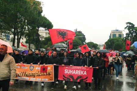 Téléchargez les photos : Tirana, Albanie - 28 novembre 2023 : Le long de l'avenue des martyrs de la nation, les gens participent joyeusement à une marche de la fête de l'indépendance un jour de pluie, tenant des drapeaux albanais aux visages souriants. - en image libre de droit
