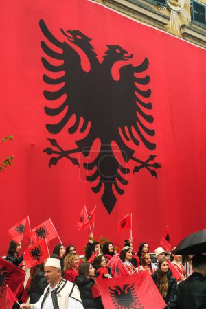 Foto de Tirana, Albania - 28 de noviembre de 2023: En un lluvioso Día de la Independencia, el Ayuntamiento de Tirana está cubierto con una enorme bandera albanesa. Los ciudadanos pasan por el edificio cubierto de bandera, marchando alegremente con caras sonrientes y sosteniendo banderas albanesas. - Imagen libre de derechos