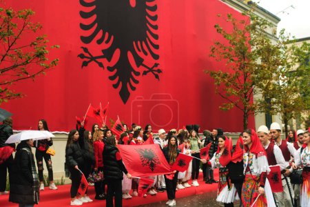 Photo for Tirana, Albania - November 28, 2023: On a rainy Independence Day, Tirana City Hall is covered with a massive Albanian flag. Citizens pass by the flag-draped building, marching joyfully with smiling faces and holding Albanian flags. - Royalty Free Image