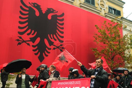 Foto de Tirana, Albania - 28 de noviembre de 2023: En un lluvioso Día de la Independencia, el Ayuntamiento de Tirana está cubierto con una enorme bandera albanesa. Los ciudadanos pasan por el edificio cubierto de bandera, marchando alegremente con caras sonrientes y sosteniendo banderas albanesas. - Imagen libre de derechos