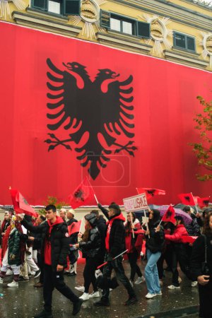 Photo for Tirana, Albania - November 28, 2023: On a rainy Independence Day, Tirana City Hall is covered with a massive Albanian flag. Citizens pass by the flag-draped building, marching joyfully with smiling faces and holding Albanian flags. - Royalty Free Image