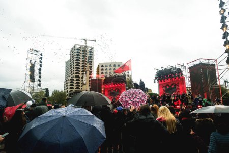 Foto de Tirana, Albania - 28 de noviembre de 2023: En la plaza Skanderbeg, en un día lluvioso de la independencia, una mujer vestida con trajes tradicionales albaneses baila jubilosamente con una bandera albanesa, con las armas extendidas, mientras una multitud con sombrillas observa, con el Mus de la Historia Nacional - Imagen libre de derechos