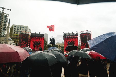 Photo for Tirana, Albania - November 28, 2023: In Skanderbeg Square on a rainy Independence Day, a woman in traditional Albanian attire dances jubilantly with an Albanian flag, arms outstretched, as a crowd with umbrellas watches, with the National History Mus - Royalty Free Image