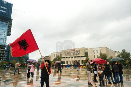 Téléchargez les photos : Tirana, Albanie - 28 novembre 2023 : Par un jour pluvieux de l'indépendance sur la place Skanderbeg, un homme se tient devant le Musée national d'histoire de Tirana, agitant un grand drapeau albanais - en image libre de droit