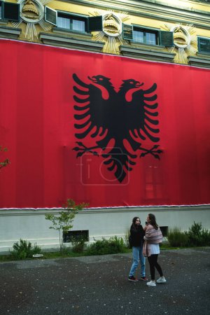 Foto de Tirana, Albania - 28 de noviembre de 2023: En un lluvioso Día de la Independencia, la muralla del Ayuntamiento de Tirana está completamente cubierta con una gran bandera albanesa, con dos jóvenes frente a ella. - Imagen libre de derechos