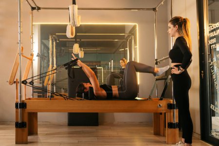Photo for Photo of a trainer instructing a member on a Reformer machine in a gym's Reformer room, capturing a focused fitness session - Royalty Free Image