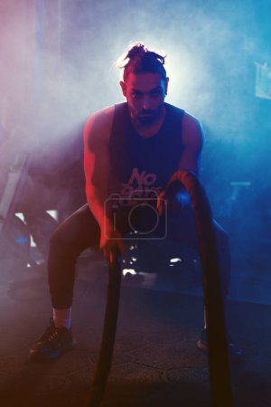Foto de Un atleta balancea cuerdas en un gimnasio bajo luces azules y rojas con un ambiente nebuloso y retroiluminado que mejora la intensidad - Imagen libre de derechos