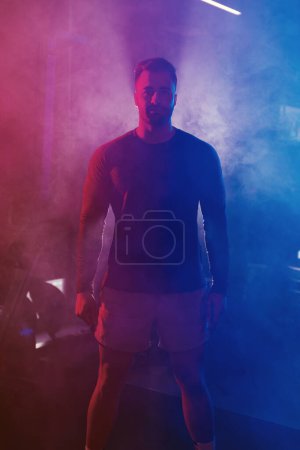 Téléchargez les photos : Un athlète masculin s'affirme, les bras sur les côtés, dans une salle de sport sous un éclairage bleu et rouge lunaire avec une atmosphère rétro-éclairée et brumeuse - en image libre de droit