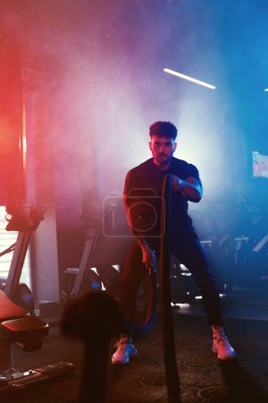 Téléchargez les photos : Un athlète balance des cordes dans une salle de gym sous des lumières bleues et rouges avec un contre-jour, réglage brumeux améliorant l'intensité - en image libre de droit