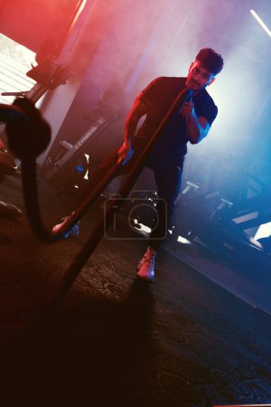 Foto de Un atleta balancea cuerdas en un gimnasio bajo luces azules y rojas con un ambiente nebuloso y retroiluminado que mejora la intensidad - Imagen libre de derechos