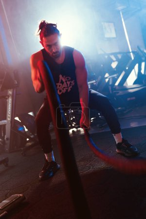 Téléchargez les photos : Un athlète balance des cordes dans une salle de gym sous des lumières bleues et rouges avec un contre-jour, réglage brumeux améliorant l'intensité - en image libre de droit