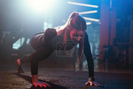 Foto de Una mujer atlética muestra flexibilidad durante un push-up, con el pie extendido sobre el hombro, bajo una dramática iluminación de gimnasio - Imagen libre de derechos