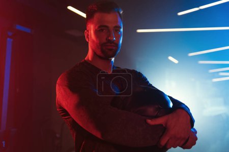 Photo for Portrait of a male athlete holding a medicine ball, captured under the neon blue and red lights of a misty gym - Royalty Free Image