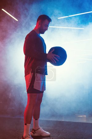 Foto de Un intenso momento de entrenamiento mientras un atleta masculino golpea una pelota de medicina contra el suelo, rodeado por un nebuloso resplandor de luces rojas y azules - Imagen libre de derechos