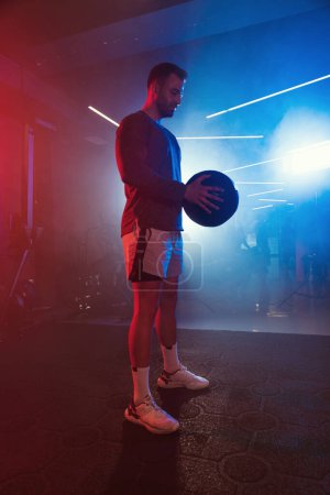 Téléchargez les photos : Portrait d'un athlète masculin tenant un ballon de médecine, capturé sous les lumières bleu néon et rouge d'un gymnase brumeux - en image libre de droit