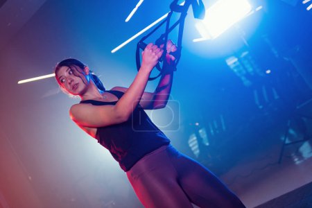 Foto de Capturada en un gimnasio, una mujer entrena con TRX sobre un telón de fondo de luces azules y rojas, creando una silueta misteriosa en la niebla - Imagen libre de derechos