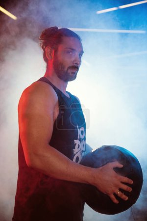 Téléchargez les photos : Portrait d'un athlète masculin tenant un ballon de médecine, capturé sous les lumières bleu néon et rouge d'un gymnase brumeux - en image libre de droit