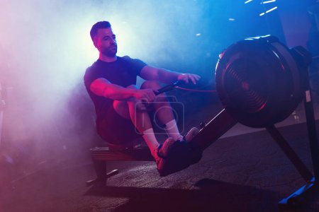 A male athlete pulls the oars on a rowing machine, his form lit by moody blue and red lights, shrouded in light fog