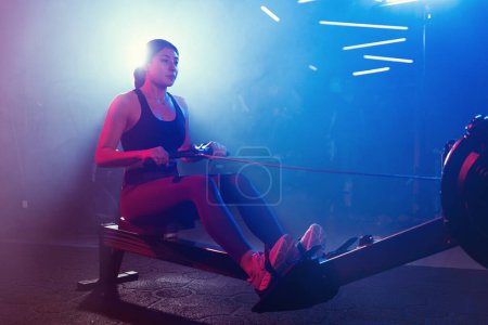 Foto de En el gimnasio, una mujer se entrena en una máquina de remo, rodeada por una mezcla mística de luces azules y rojas y un toque de niebla - Imagen libre de derechos