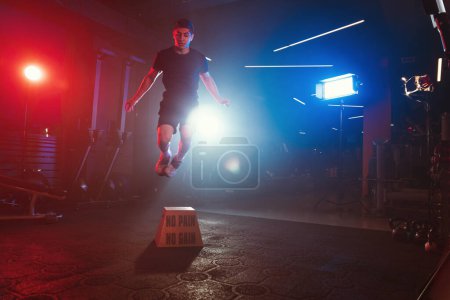 Téléchargez les photos : Un athlète exécute un saut en boîte, s'élevant au-dessus de "No Pain No Gain", au milieu d'une salle de sport illuminée par des lumières bleues et rouges et une brume molle - en image libre de droit