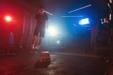 Téléchargez les photos : Un athlète exécute un saut en boîte, s'élevant au-dessus de "No Pain No Gain", au milieu d'une salle de sport illuminée par des lumières bleues et rouges et une brume molle - en image libre de droit