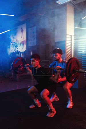Téléchargez les photos : Un athlète masculin concentré exécute des squats sous la direction d'un entraîneur, mis en évidence par les lumières vives du gymnase bleu et rouge au milieu d'une brume subtile - en image libre de droit