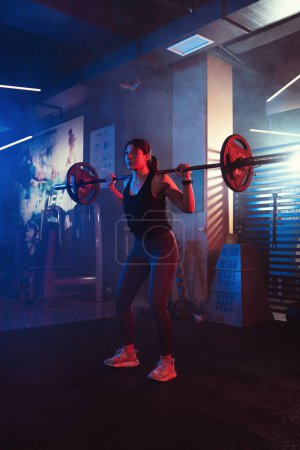 Unter den wachsamen Augen ihres Trainers hockt eine Athletin mit einer Langhantel in einem von stimmungsvollen blauen und roten Lichtern erleuchteten Fitnessstudio