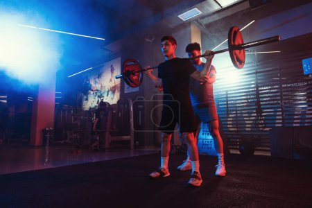 A focused male athlete performs squats under the guidance of a trainer, highlighted by the vivid blue and red gym lights amidst a subtle mist