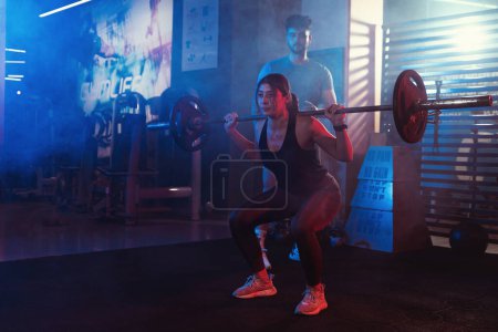 Foto de Bajo la atenta mirada de su entrenador, una atleta se agacha con una barra en un gimnasio iluminado por las luces azules y rojas atmosféricas - Imagen libre de derechos