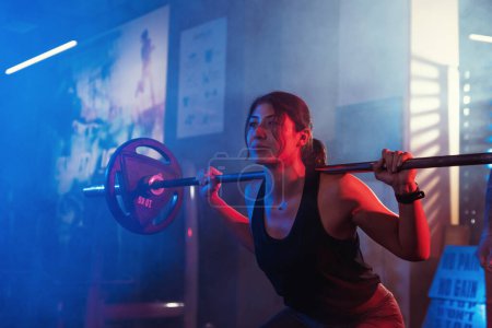 Bajo la atenta mirada de su entrenador, una atleta se agacha con una barra en un gimnasio iluminado por las luces azules y rojas atmosféricas