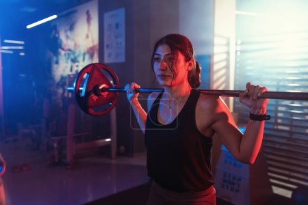 Under the watchful eye of her trainer, a female athlete squats with a barbell in a gym lit by atmospheric blue and red lights