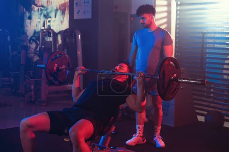 Focused male athlete performing an incline bench press under the watchful eye of his trainer, illuminated by vibrant blue and red gym lights amidst a misty ambiance
