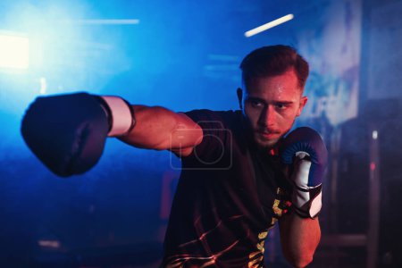 Foto de Boxeador masculino enfocado practica golpes solo en un gimnasio con dramática iluminación roja y azul, creando una atmósfera viva y enérgica - Imagen libre de derechos
