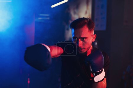 Téléchargez les photos : Le boxeur masculin concentré pratique les coups de poing seul dans une salle de gym avec un éclairage rouge et bleu dramatique, créant une atmosphère vive et énergique - en image libre de droit