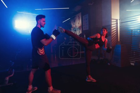 A woman executes a high kick as her partner defends in a gym session under moody lightin