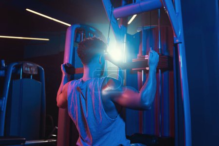 Photo for A man perfects his form on the lat pull down machine, a fusion of focus and strength captured amidst the gym's mystic blue and red hues - Royalty Free Image
