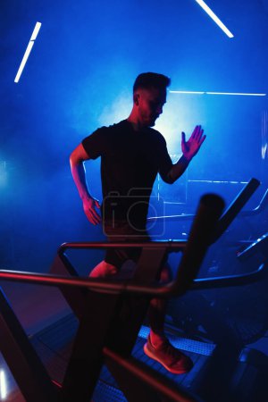 Téléchargez les photos : Athlète masculin courant sur un tapis roulant, illuminé par des lumières bleues et rouges vives créant un environnement de gymnase atmosphérique et lunatique - en image libre de droit
