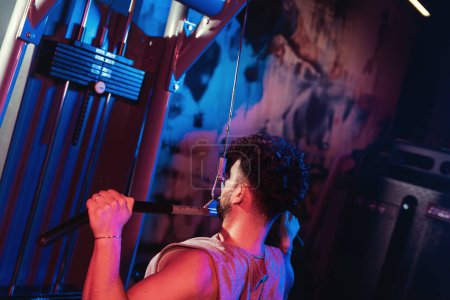 Téléchargez les photos : Un homme perfectionne sa forme sur la machine de traction latérale, une fusion de concentration et de force capturée dans les teintes mystiques bleu et rouge de la salle de gym - en image libre de droit