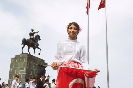 Foto de Izmir, Turquía - 23 de abril de 2024 Una niña alegre ondea la bandera turca durante las festividades del Día de los Niños con un traje tradicional, con la estatua de Ataturk a caballo en el fondo - Imagen libre de derechos