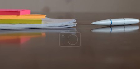 Multicolored paper stickers and white ballpoint pen close-up on wooden table with reflections on blurred background