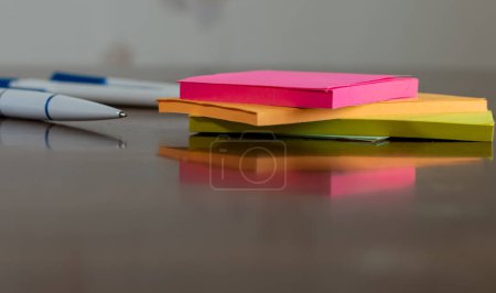 Multicolored paper stickers and white ballpoint pen close-up on wooden table with reflections on blurred background