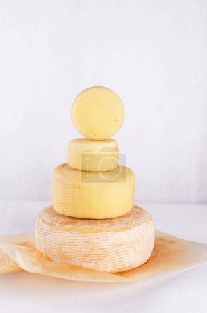 Photo for A pile of different kinds cheese heads stacked in vertical row on table. Medium hard cheese heads on wooden cutting board. Healthy organic eating concept. Handmade cheese. - Royalty Free Image