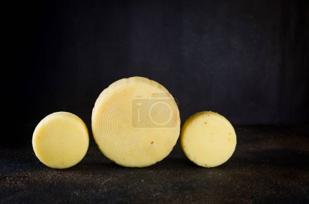 Foto de Muchos tipos diferentes cabezas de queso en la mesa oscura. Cabezas de queso duro medio sobre tabla de cortar de madera. Concepto de alimentación orgánica saludable. Queso hecho a mano. Productos lácteos frescos, - Imagen libre de derechos