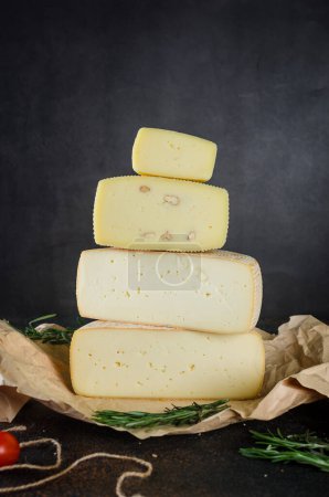 Foto de Muchos tipos diferentes cabezas de queso en fila vertical en la mesa oscura. Cabezas de queso duro medio sobre tabla de cortar de madera. Concepto de alimentación orgánica saludable. Queso hecho a mano. Productos lácteos frescos, - Imagen libre de derechos