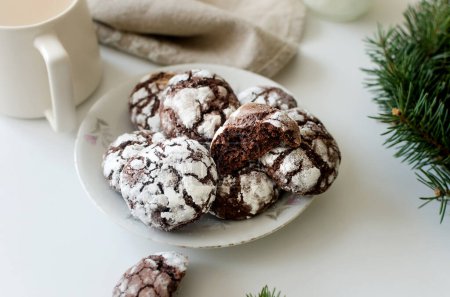 Homemade Cracked chocolate biscuits in white plate and cup tea with milk on table. Traditional American chocolate cookies for Christmas with cracks and powdered sugar 