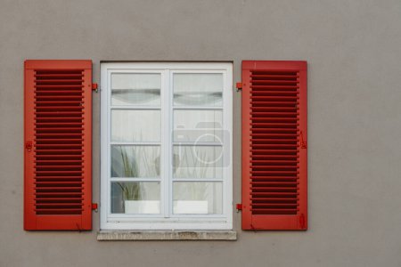 Italienische Fenster an der weißen Fassade mit offenen roten Fensterläden und Blumen an den Fenstern. Fensterladenwand