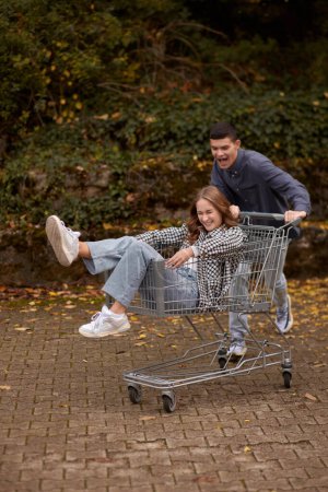 Autumn Joyride: Boy Rolling Girl in Shopping Cart amid Natures Beauty. playful couple, autumnal adventure, outdoor fun, seasonal joy, laughter and love, romantic play, natures embrace, joyful