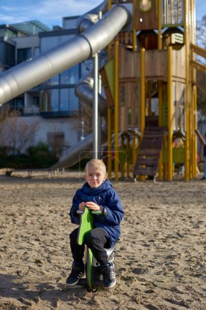 Foto de Childhood Joy: Beautiful 8-Year-Old Boy in Jacket Swinging on Horse-Shaped Seesaw, Background of Playful Park in Bietigheim-Bissingen, Germany, Autumn. Captura la esencia pura de la felicidad infantil - Imagen libre de derechos