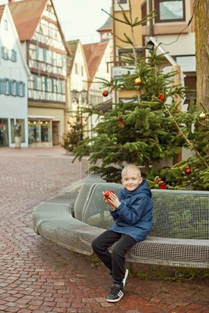Winter Wonderland Delight: 8-jähriger Junge mit Weihnachtsdekor am Vintage Fountain. Erleben Sie die Magie der Winterfreude mit diesem bezaubernden Bild, in dem ein wunderschöner 8-jähriger Junge auf einem